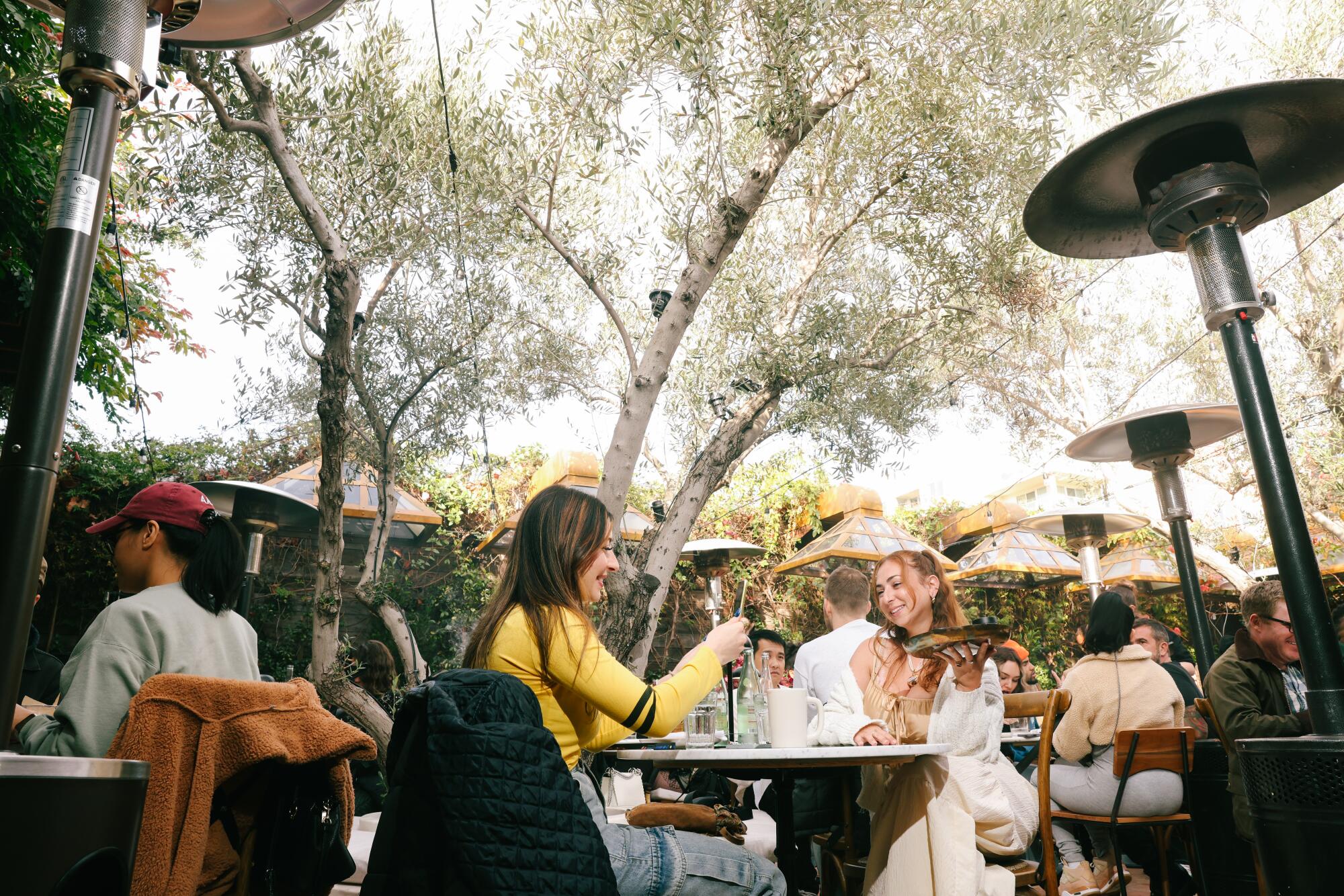 Two women sit at a table on a leafy outdoor patio, one holding up a tray and the other taking a photo with her phone.