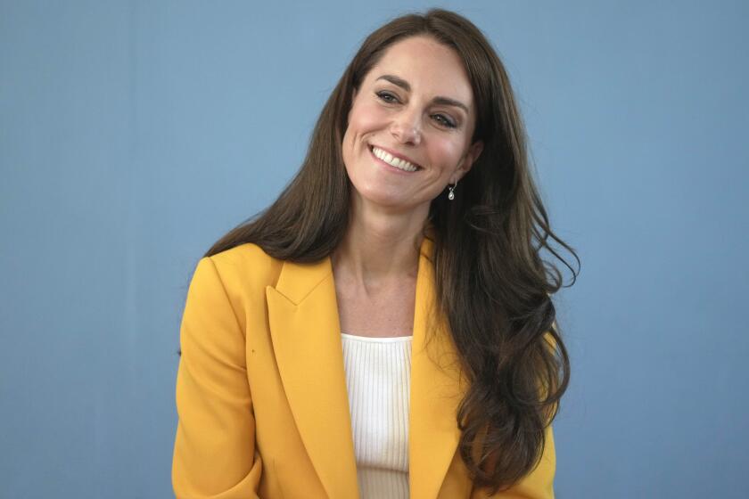 Kate Middleton in a white shirt, bright yellow blazer leaning to her left and smilling against a blue background