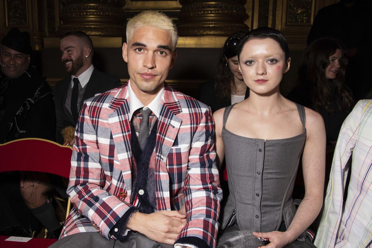 A man with platinum hair in a plaid coat sitting next to a woman with short dark hair in a gray sleeveless dress