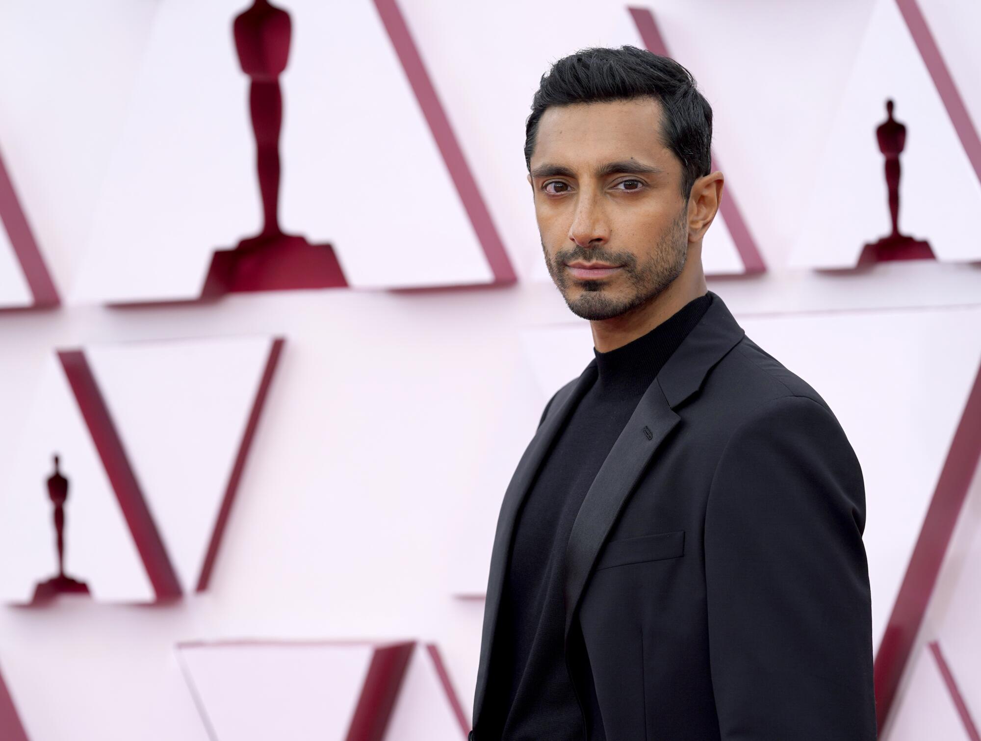 Riz Ahmed in a black suit over a black shirt.