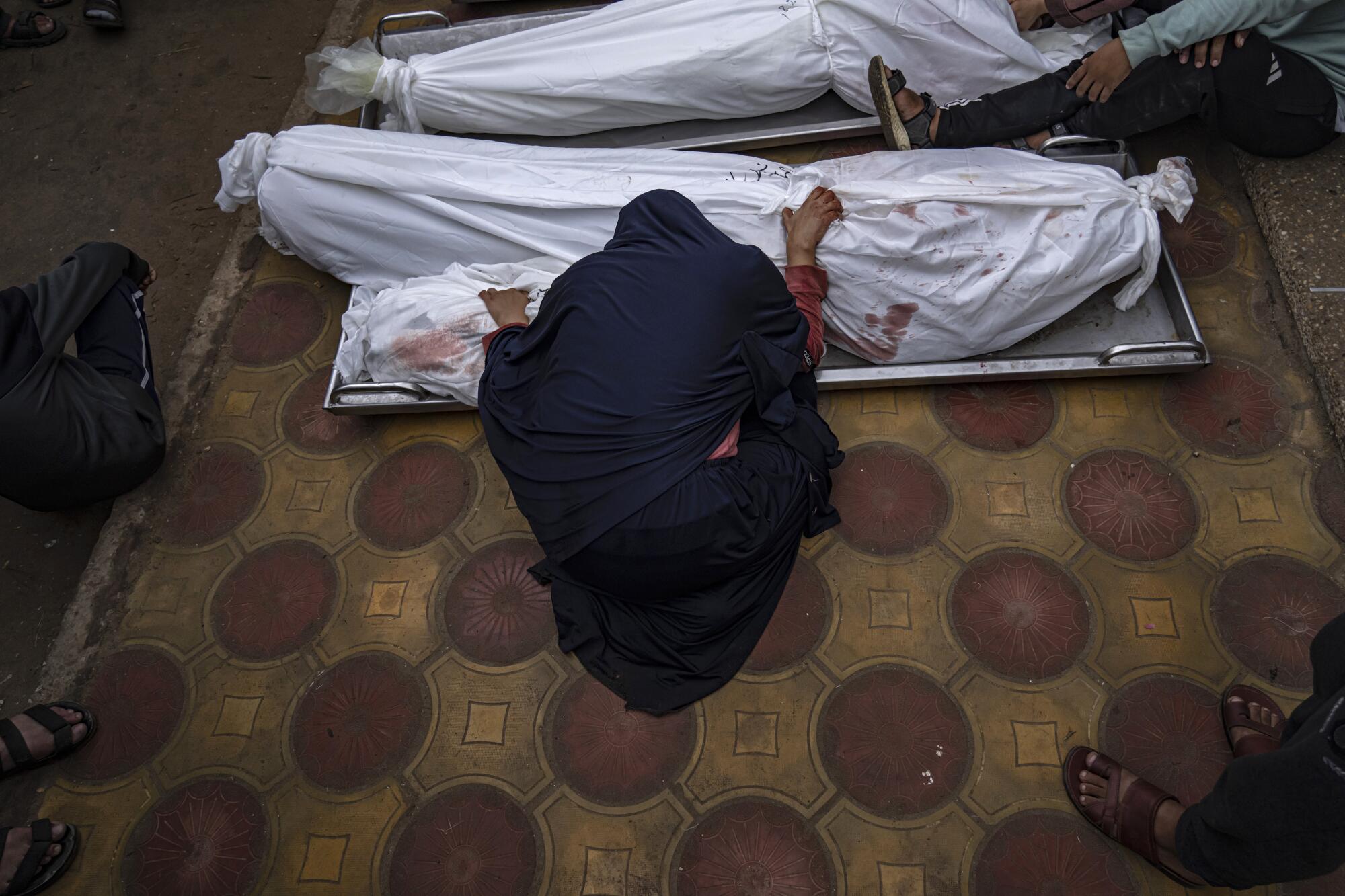 A woman in black leans over bodies shrouded in white cloth 