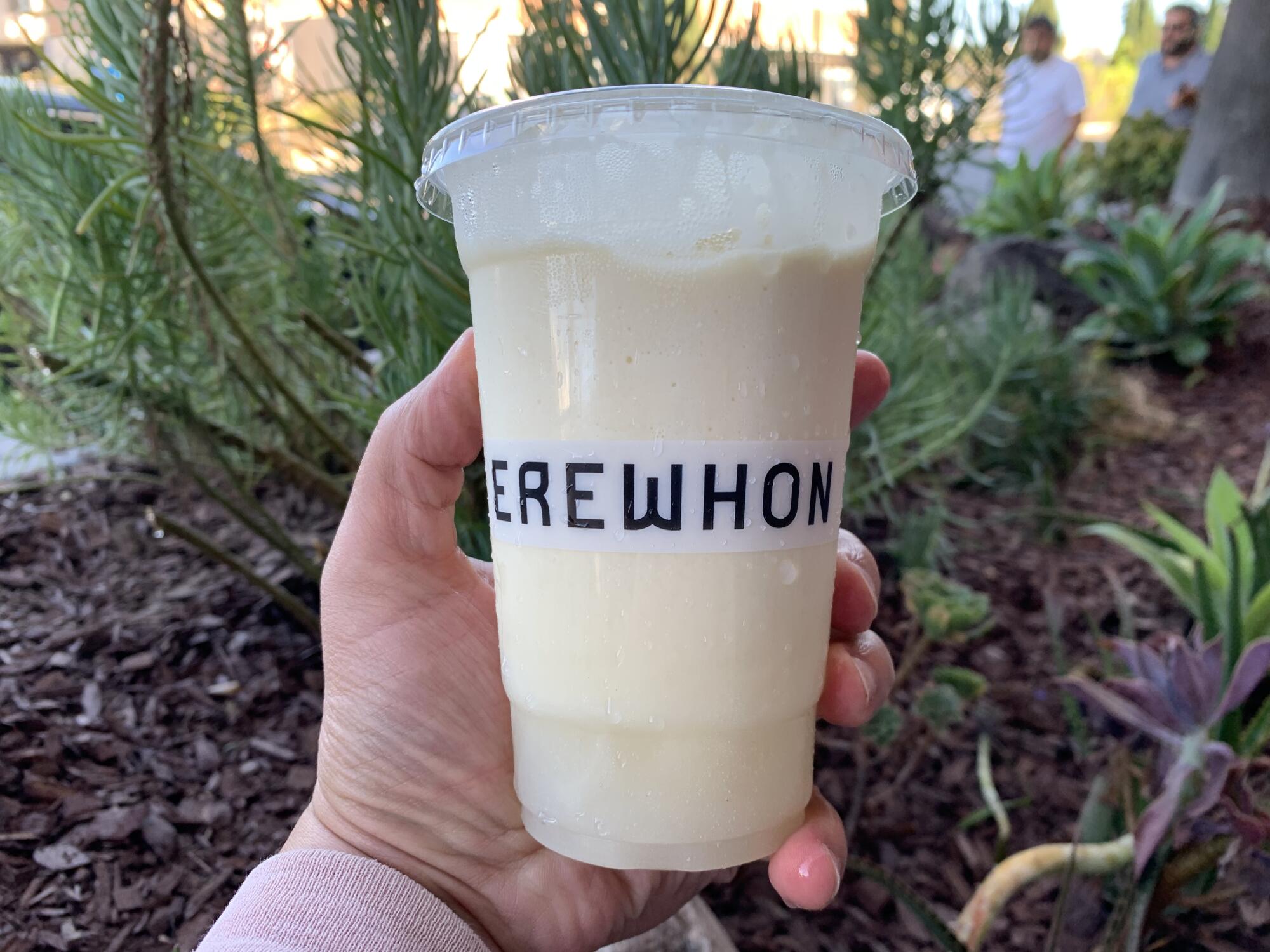 A hand holds a pale yellow smoothie in a plastic cup that says Erewhon.