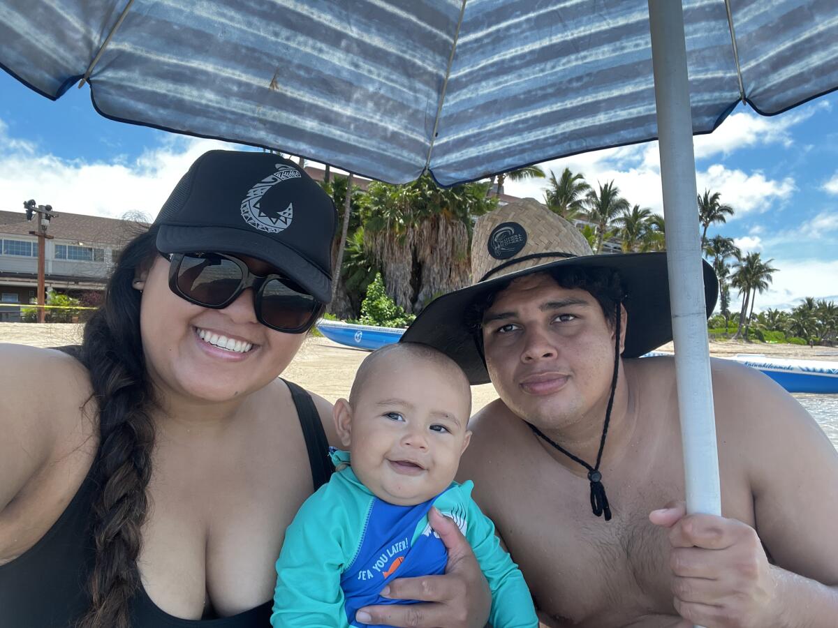 A woman in dark cap and sunglasses holds a smiling baby in a turquoise top next to a shirtless man in a hat under an umbrella