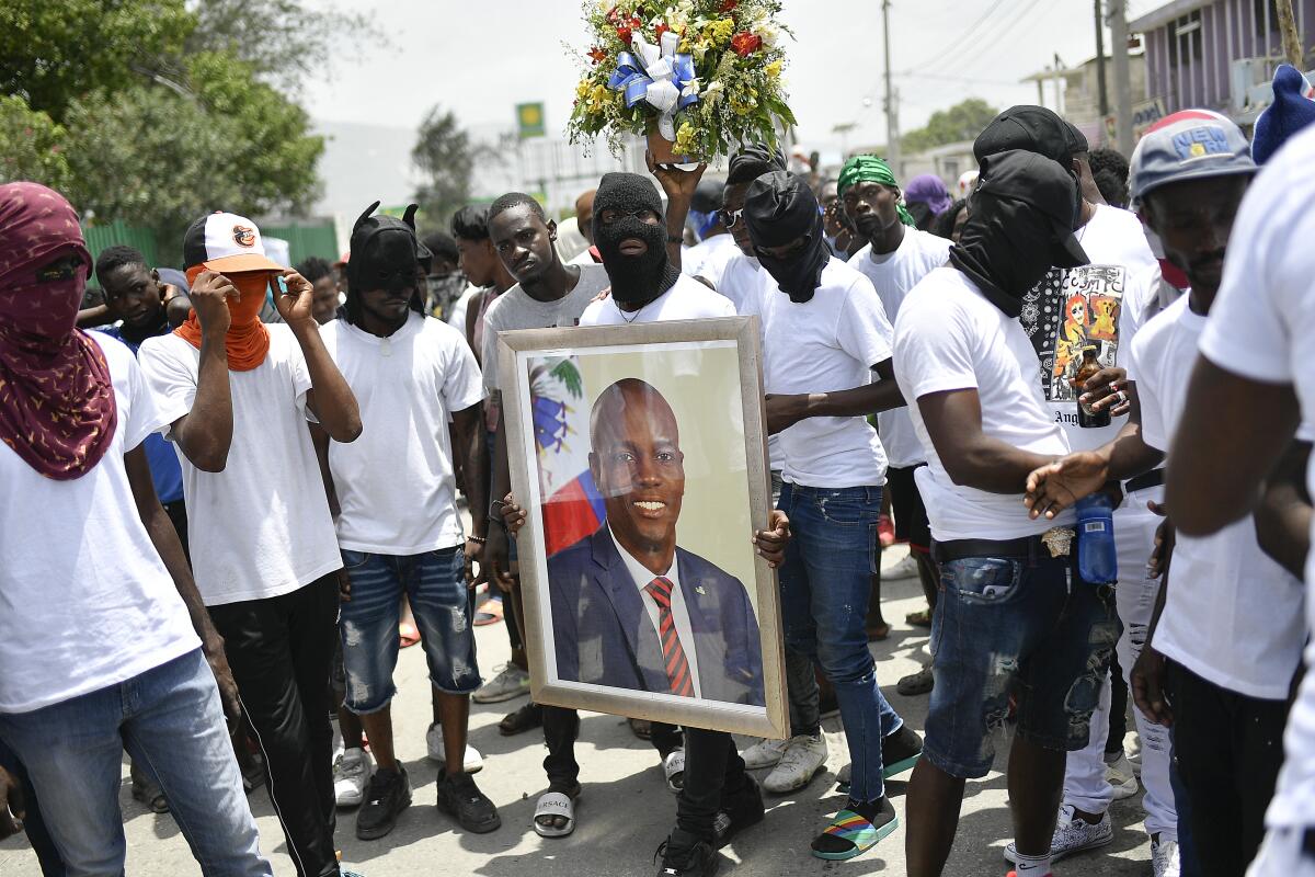 A masked man in white T-shirt and jeans, surrounded by a similarly dressed crowd, holds up a framed portrait of a man