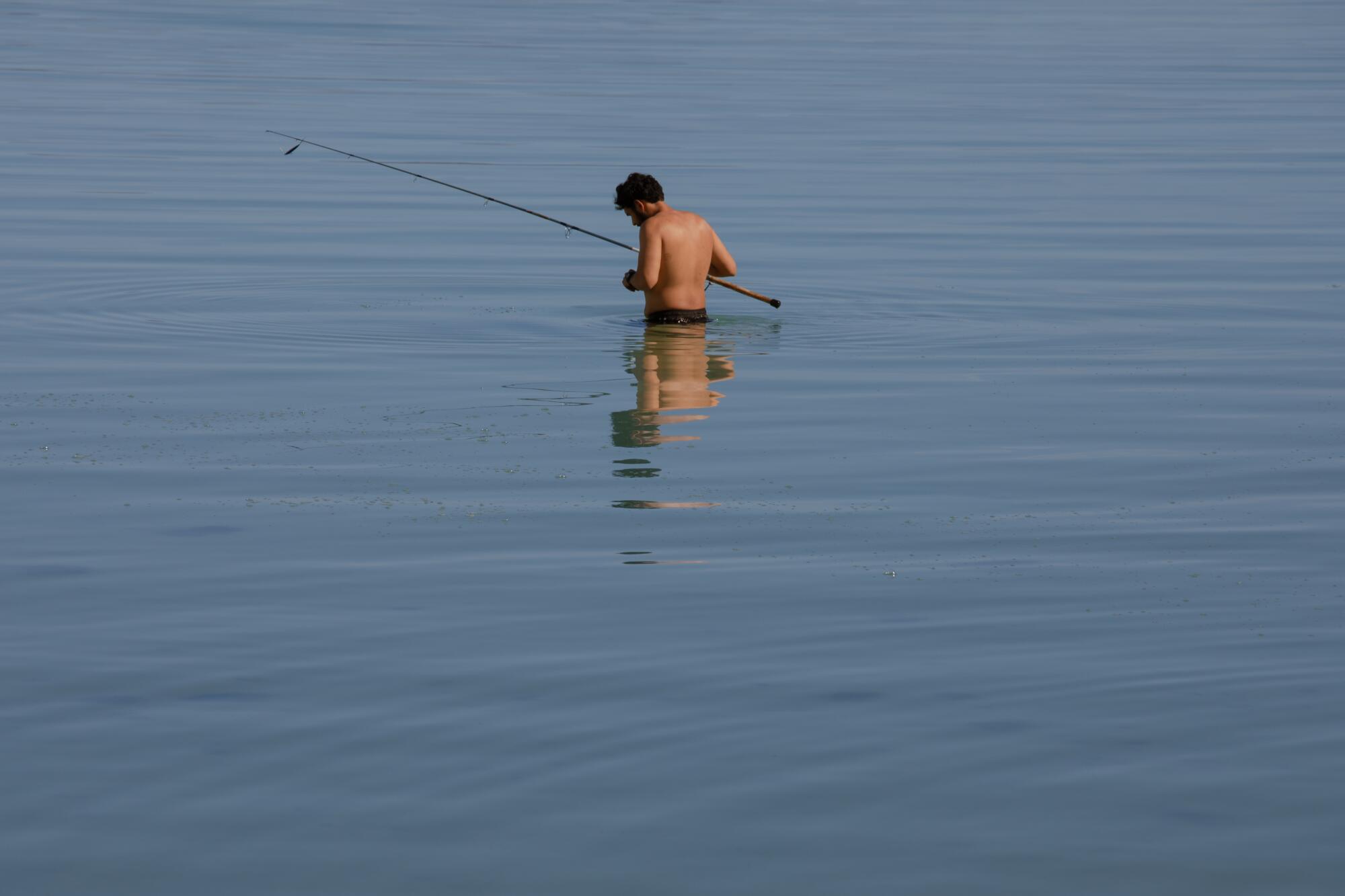 A shirtless man with a fishing rod, turned away from the camera, waist deep in and surrounded by blue water.