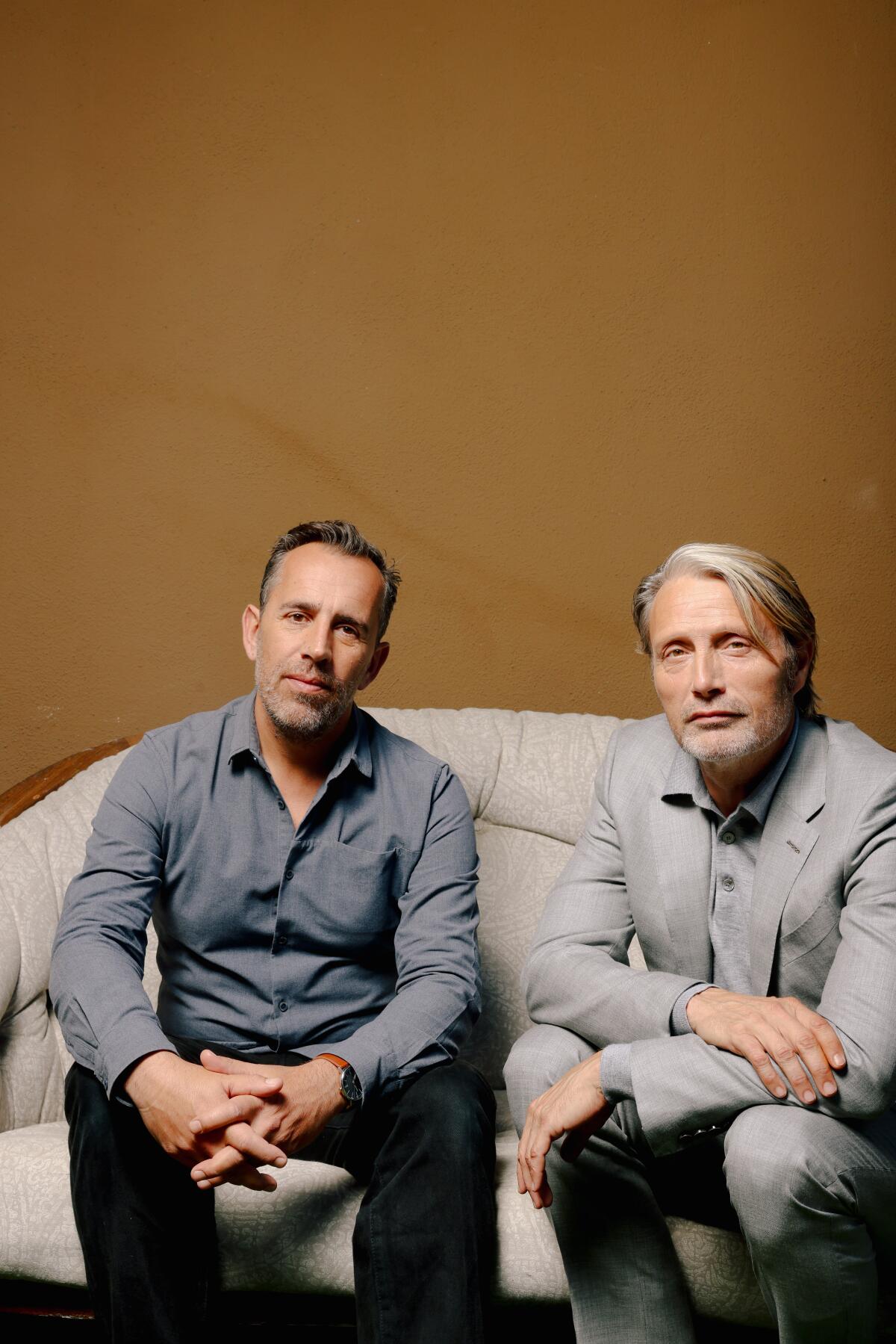 Director Nikolaj Arcel and actor Mads Mikkelsen sit on a couch and look intently at the camera.