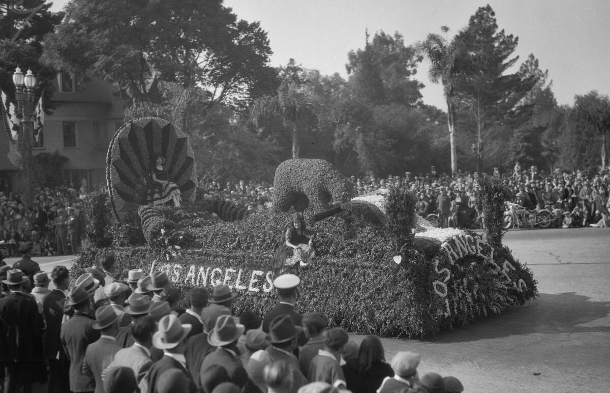 Two people ride a greenery-covered float that says "Los Angeles" on the front. The crowd of spectators is mostly in hats.