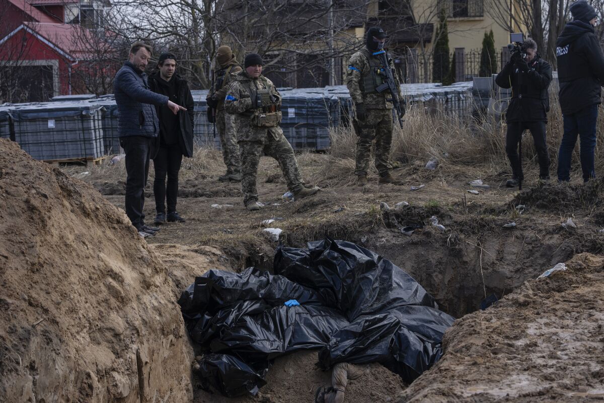People, some in fatigues, stand near a ditch with black body bags in it 