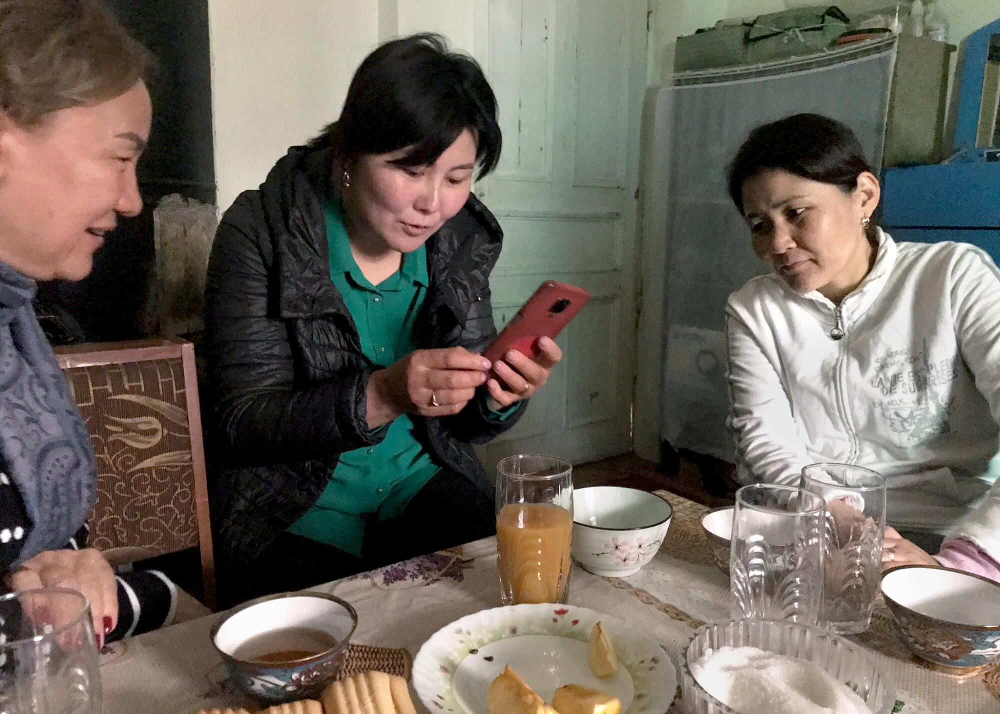 Three women sit at a kitchen table, looking at a phone, talking.