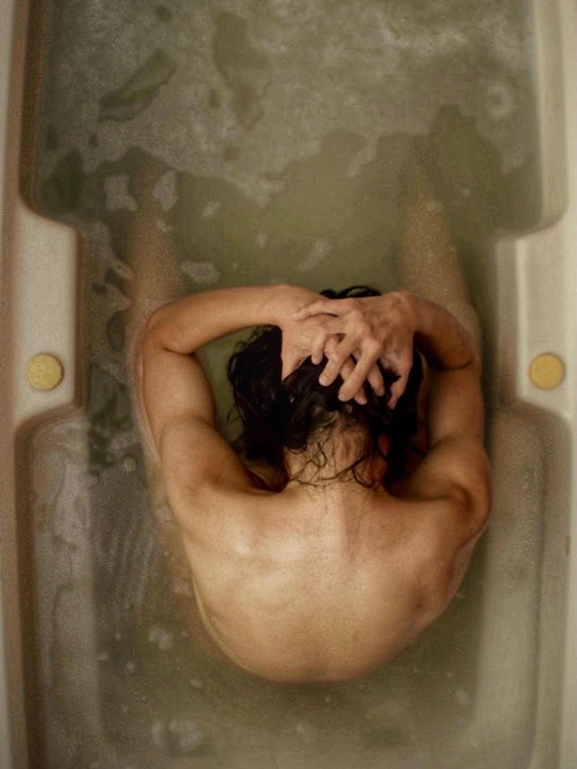 A person seen from above, sitting nude in a bathtub, hands on head.