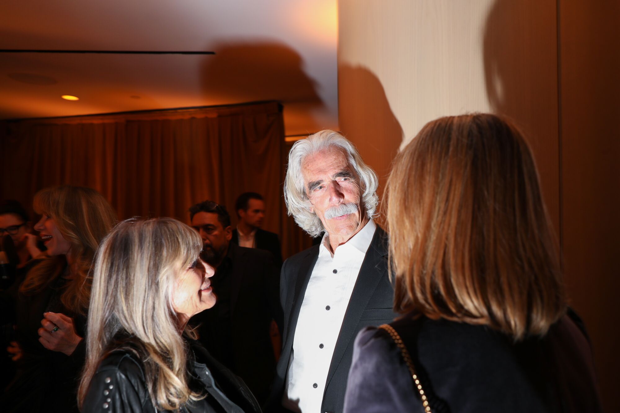 a man in a tux with a white mustache and head of hair talks to two women 