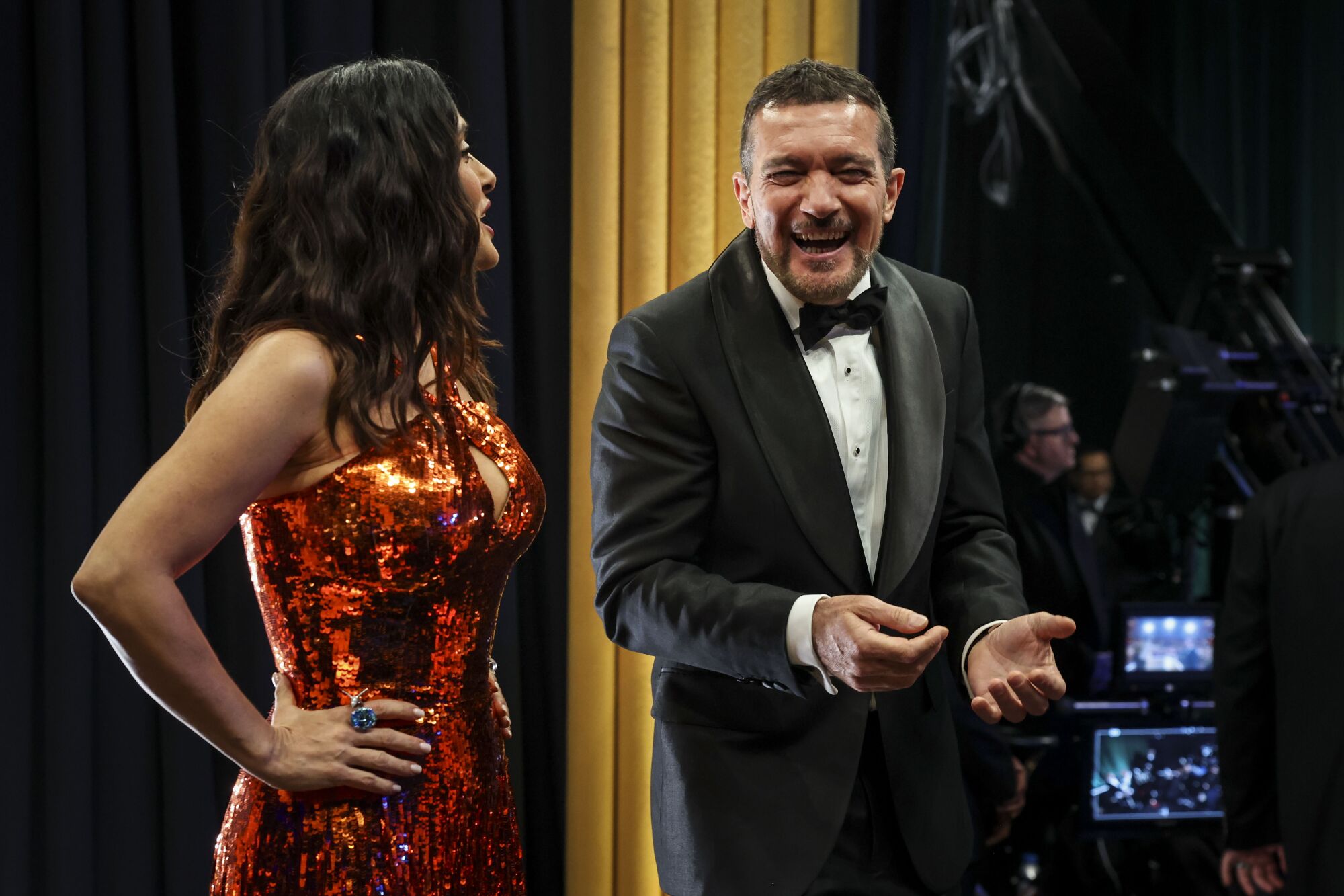 A woman in red and a man in a tux share a laugh backstage at the Oscars. 