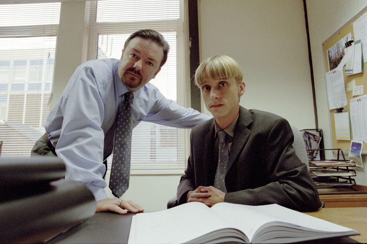 Two men, one standing and leaning on a desk, the other sitting down, look into the camera in a scene from “The Office.”