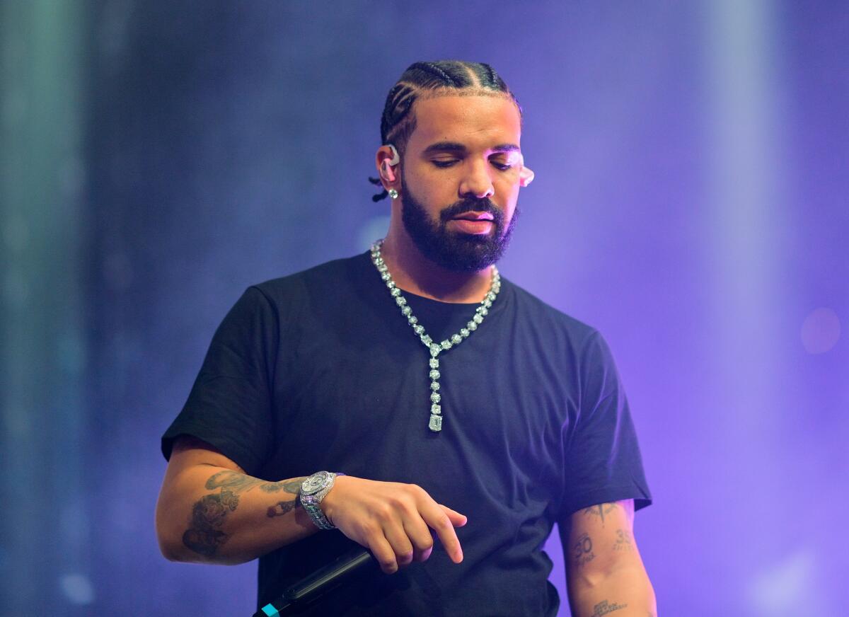 Drake, hair in cornrows, looks down on stage while wearing black T-shirt and diamond chain and holding a mic at his belly