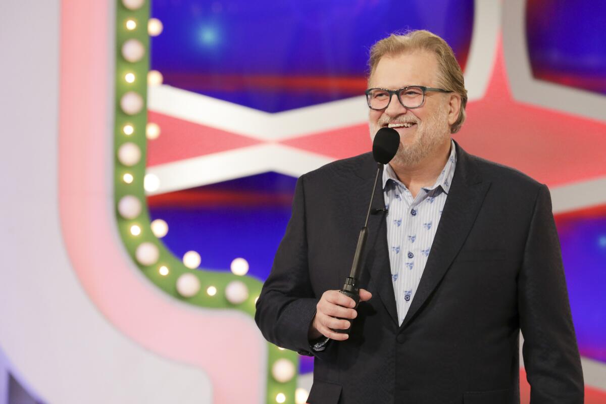 A man wearing a black suit with a white and blue shirt and no tie, smiling while holding a microphone on a TV game show. 