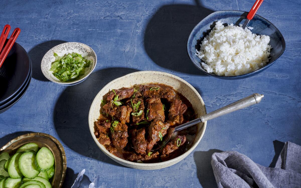 A bowl of spare ribs in a rich brown sauce, among bowls of steamed rice, sliced cucumbers and a dish of sliced scallions.