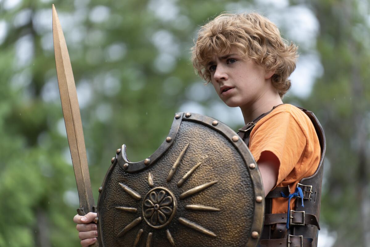 A boy with curly blond hair holding a gold shield and sword stands in the woods.