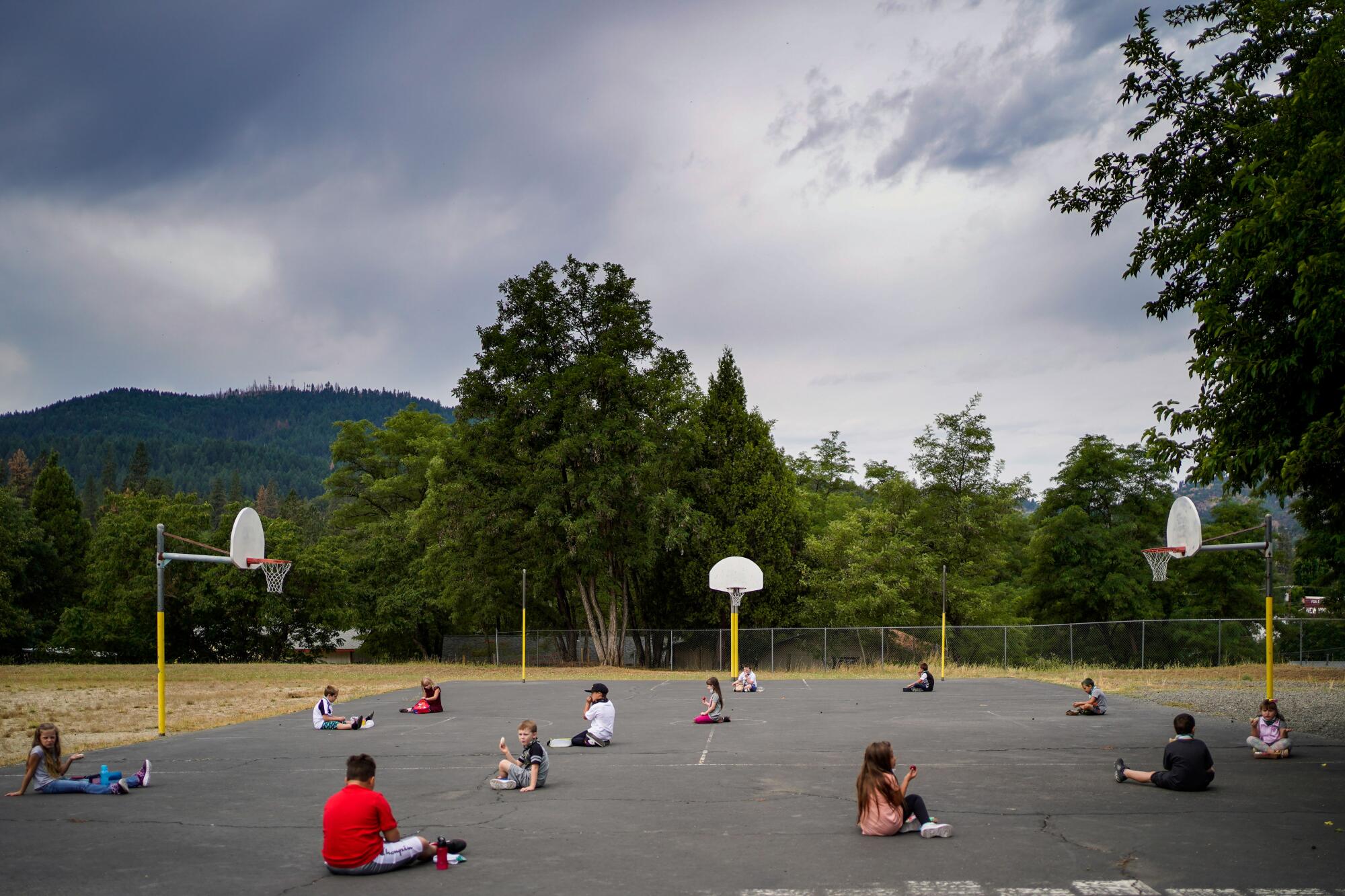 Schoolchildren eat lunch while sitting on a blacktop basketball court surrounded by greenery and forested hills.