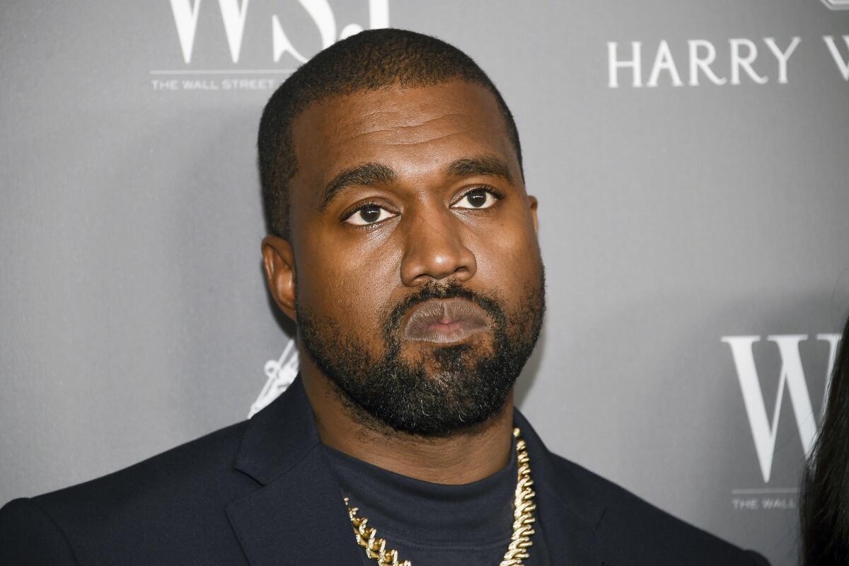 Kanye West wearing a black blazer and gold chain posting against a gray background