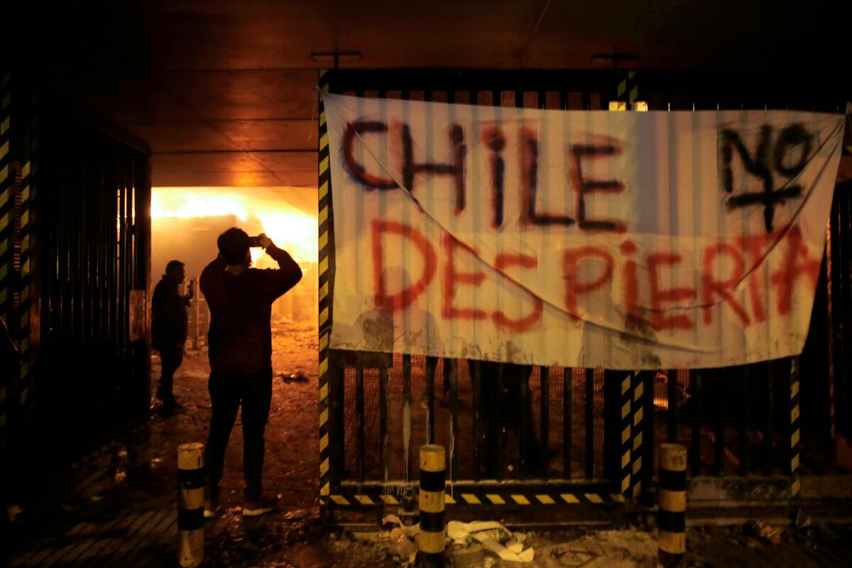 People take pictures of Macul Metro station set on fire by protesters alongside a sign that reads "Chile doesn't wake up" during a mass fare-dodging protest in Santiago, on October 19, 2019. - The entire Santiago Metro, which mobilizes about three million passengers per day, stopped operating on Friday afternoon following attacks in rejection of the rate hike, the company said. The chaos beat Santiago this Friday with confrontations, fires and attacks on the metropolitan railway, in protest of the increase in fares that forced the closure of all Metro stations. (Photo by JAVIER TORRES / AFP) (Photo by JAVIER TORRES/AFP via Getty Images) ** OUTS - ELSENT, FPG, CM - OUTS * NM, PH, VA if sourced by CT, LA or MoD **