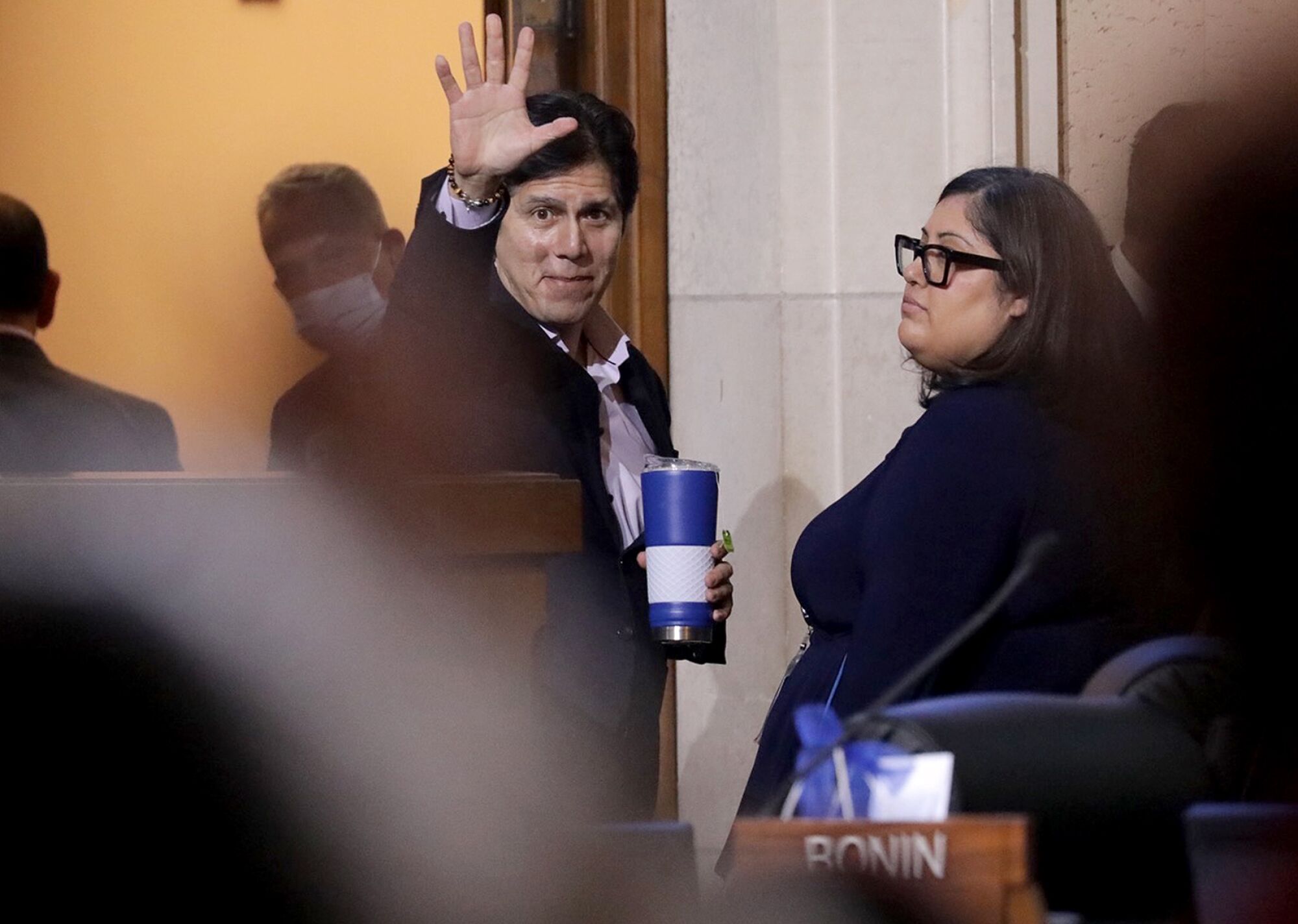 Councilor Kevin de León waves during a council meeting while other people look on.
