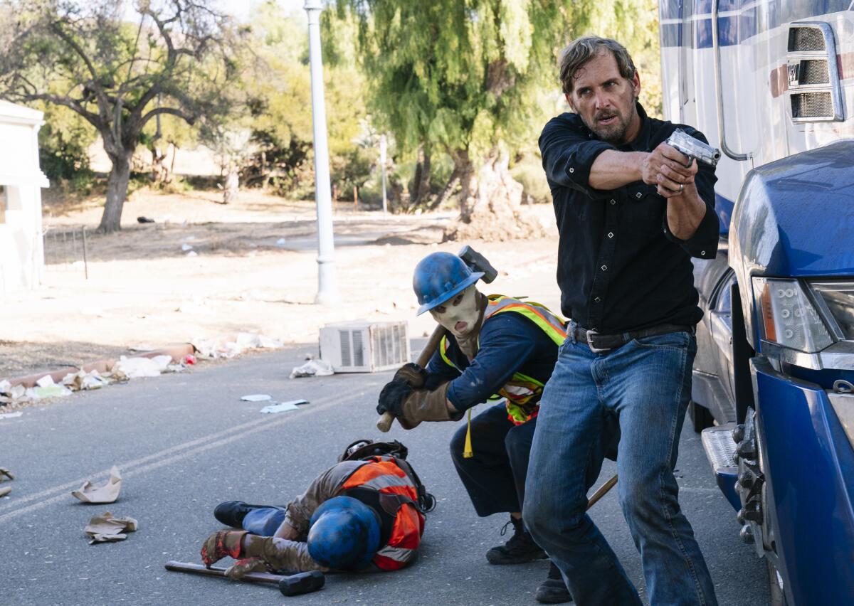 A man points a gun as a masked man squats behind him holding a sledgehammer.