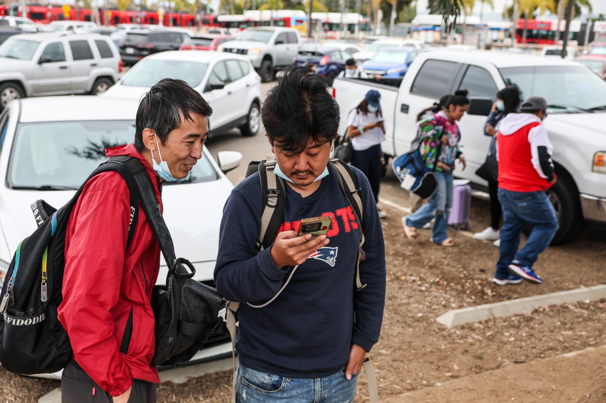 A man using his phone as another looks on and other people walk behind them in a dirt parking lot