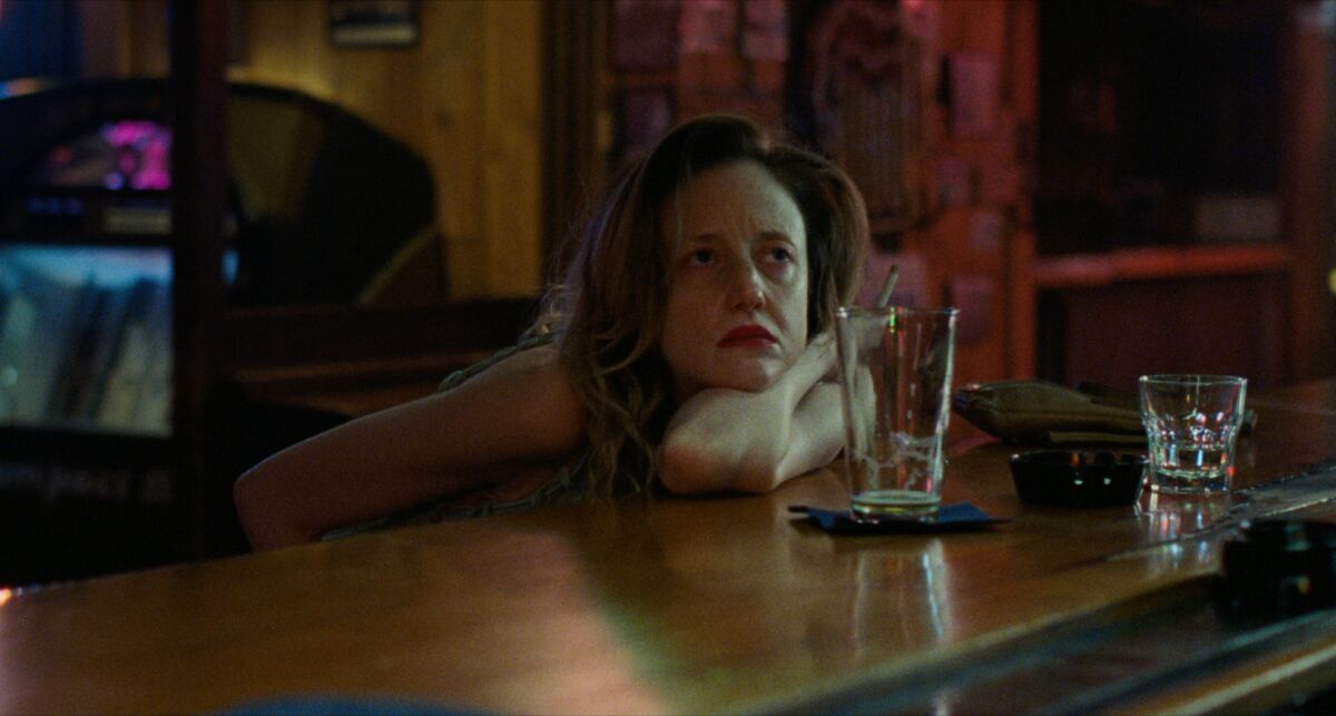 A woman holding a cigarette leans on a bar counter, empty beer glass in front of her in a scene from "To Leslie."