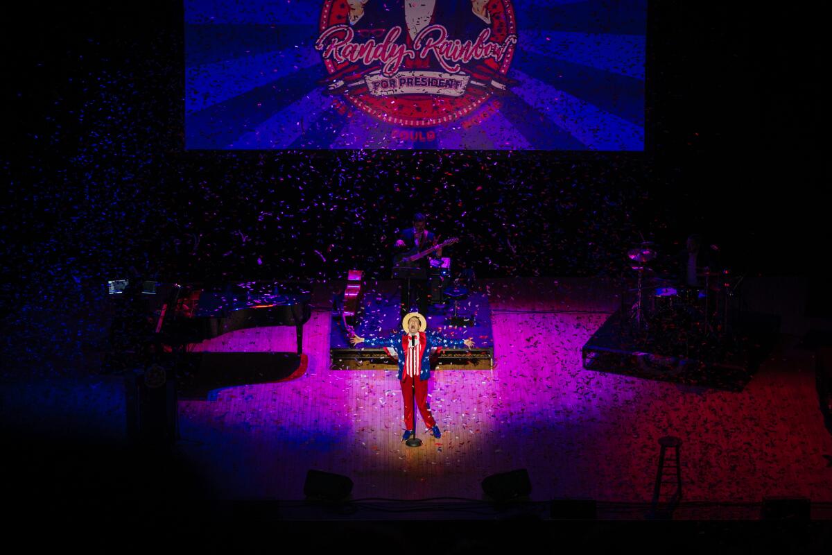 Performer Randy Rainbow, wearing red, white and blue, is seen standing on a broad stage illuminated in pink.