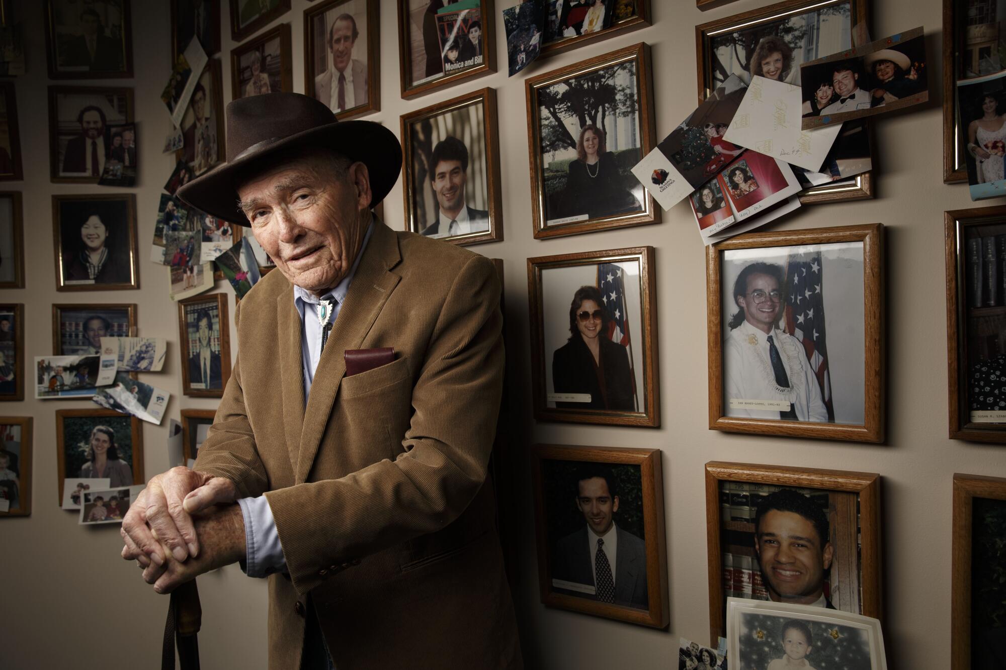 Judge Harry Pregerson, in a brown hat and blazer, stands with his hands on a cane, dozens of portraits on the wall behind him