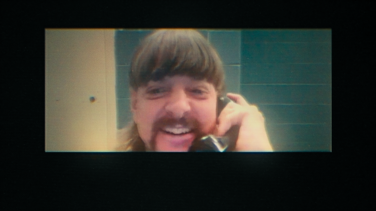 A grainy image of a man smiling while talking into a prison telephone