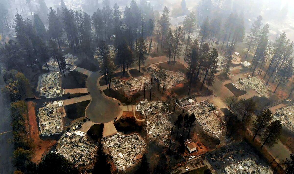 Aerial view of a block of destroyed homes and charred trees with smoke lingering in the sky