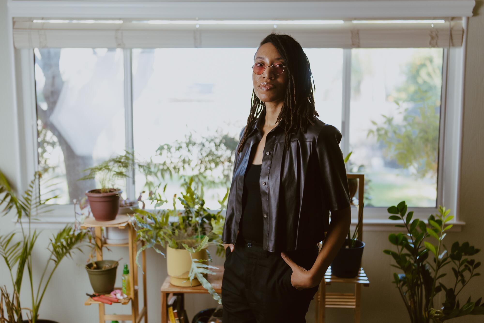 Venita Blackburn, a Black woman with locs and rose-colroed glasses stands before window and shelf full of plants.