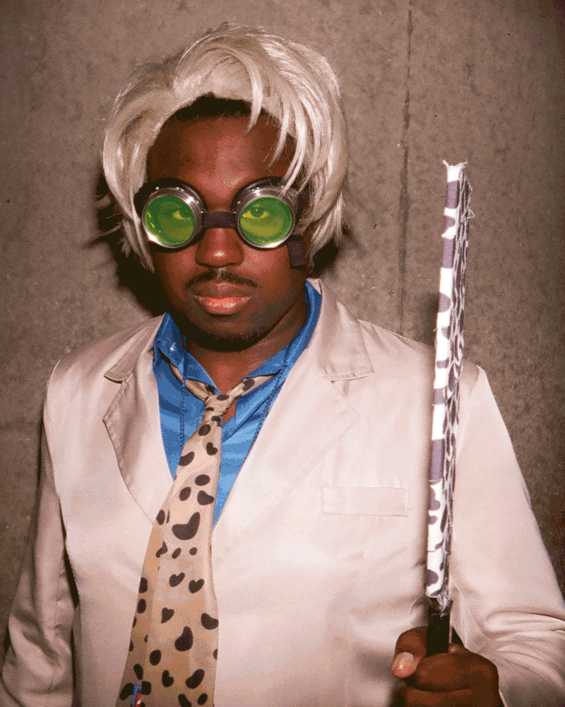 A person wearing a blond wig, green goggles, a spotted tie, and a light jacket over a blue shirt.