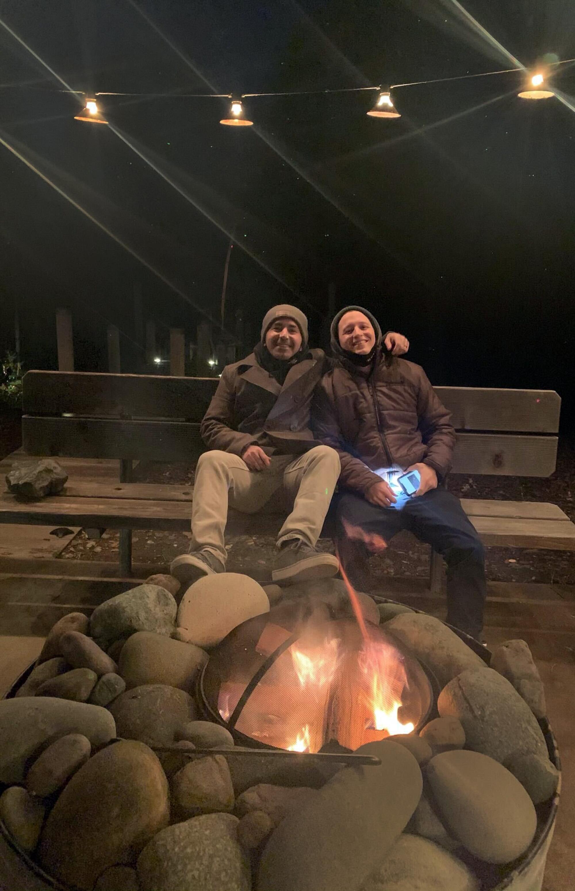Two people sit behind a fire outdoors.