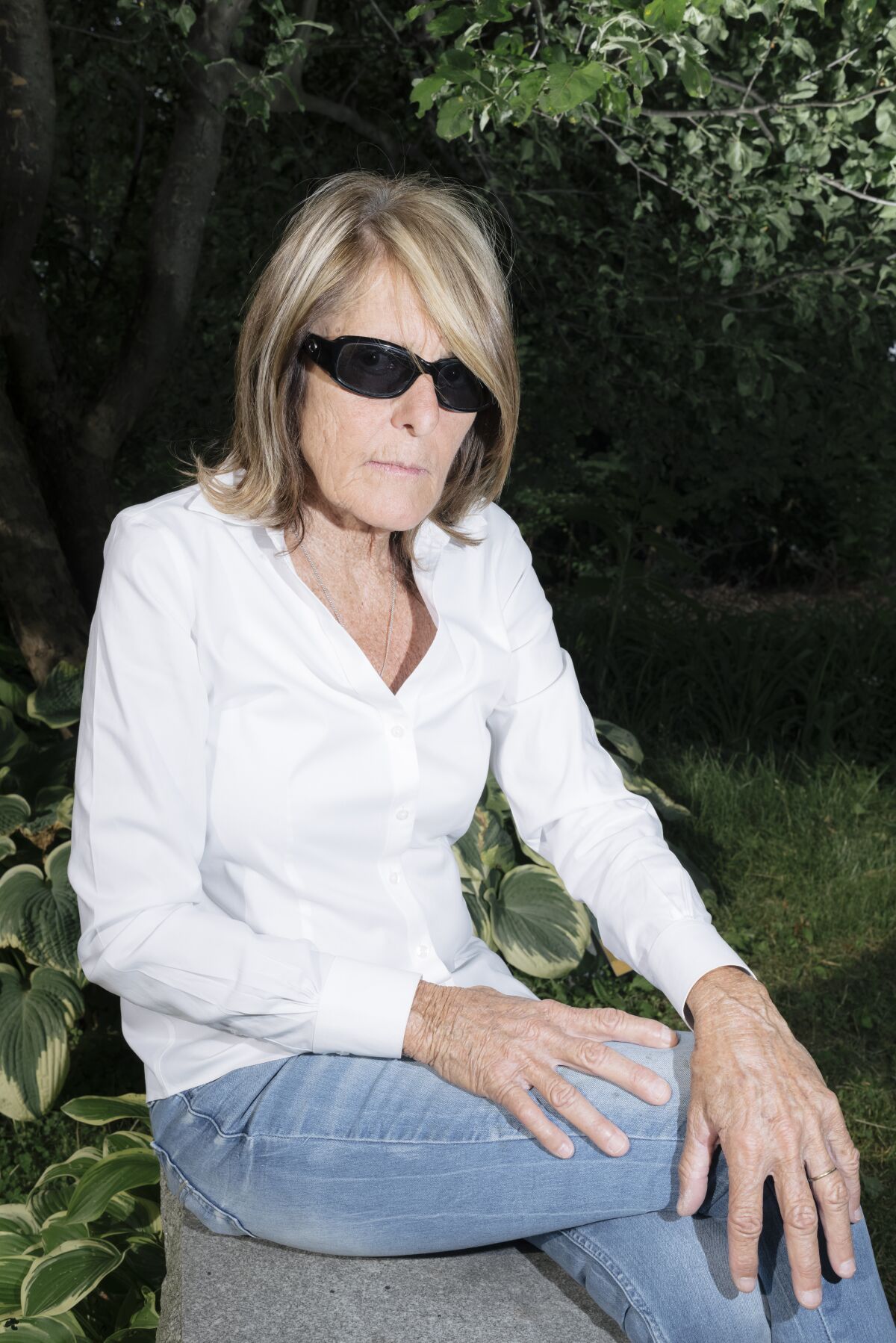 A woman wearing sunglasses sits on a rock with her legs crossed.
