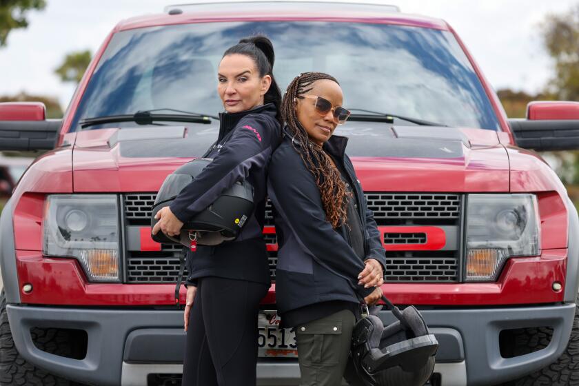 Two women pose back to back while carrying helmets in front of a red Ford truck.