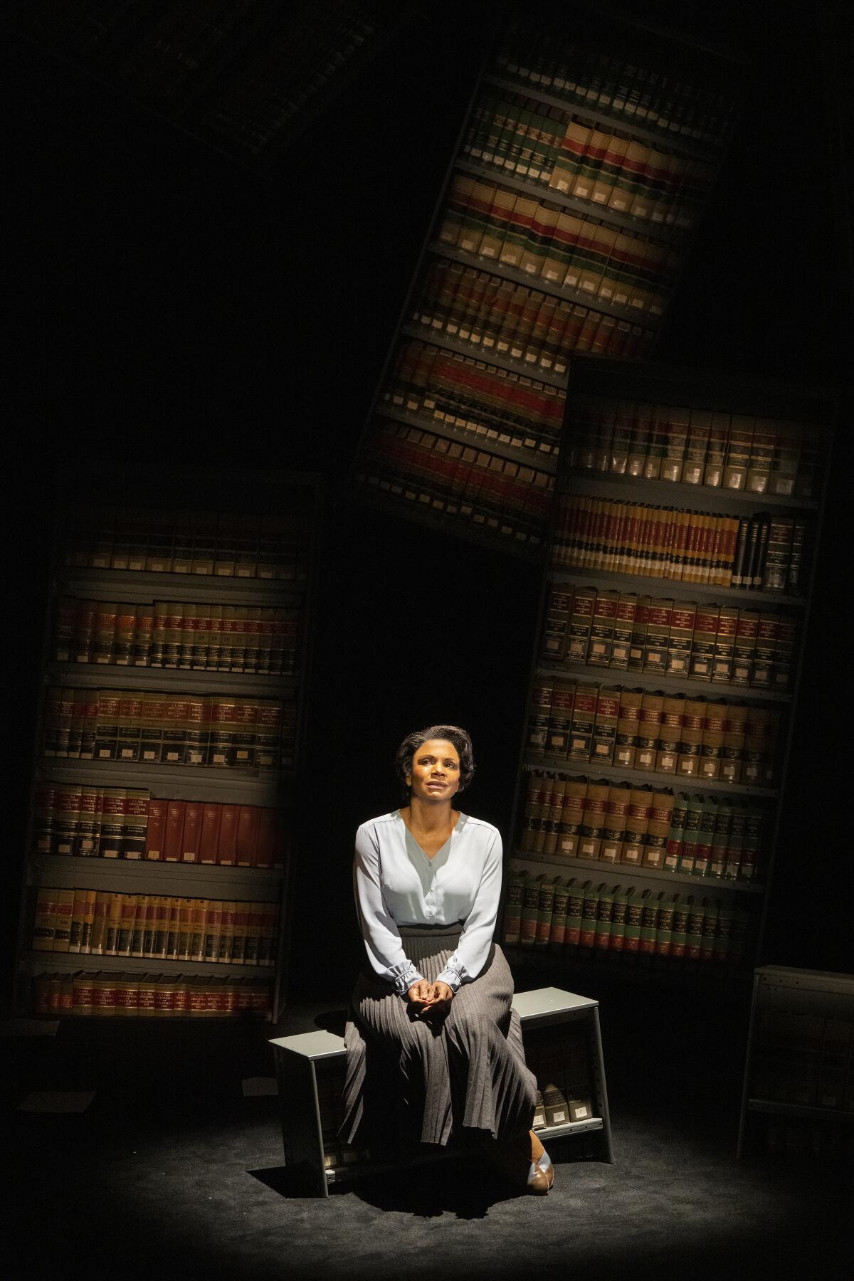 An actor sits onstage in front of bookshelves.