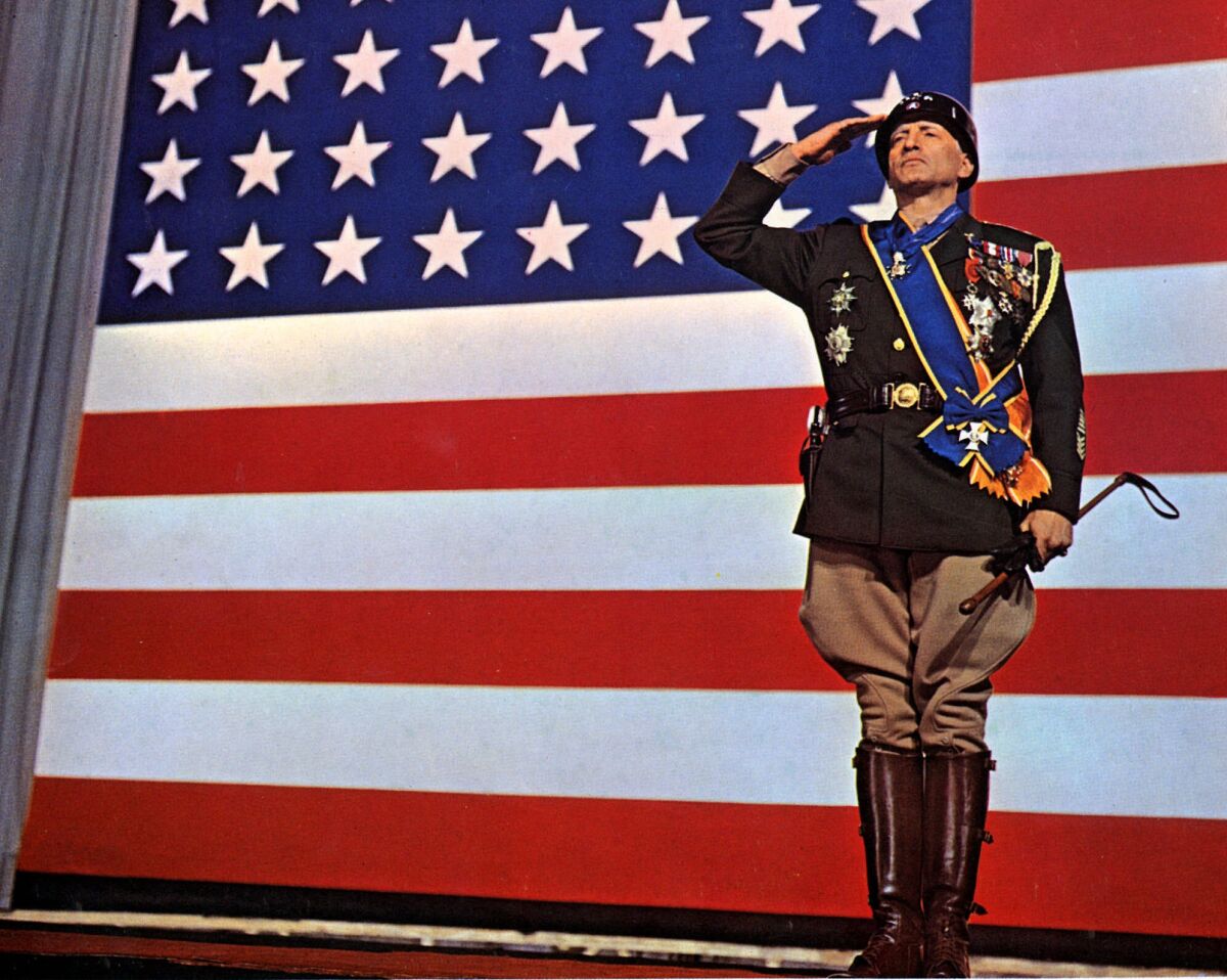 A man in a general's uniform festooned with medals, ribbons and sashes salutes with a giant American flag behind him.