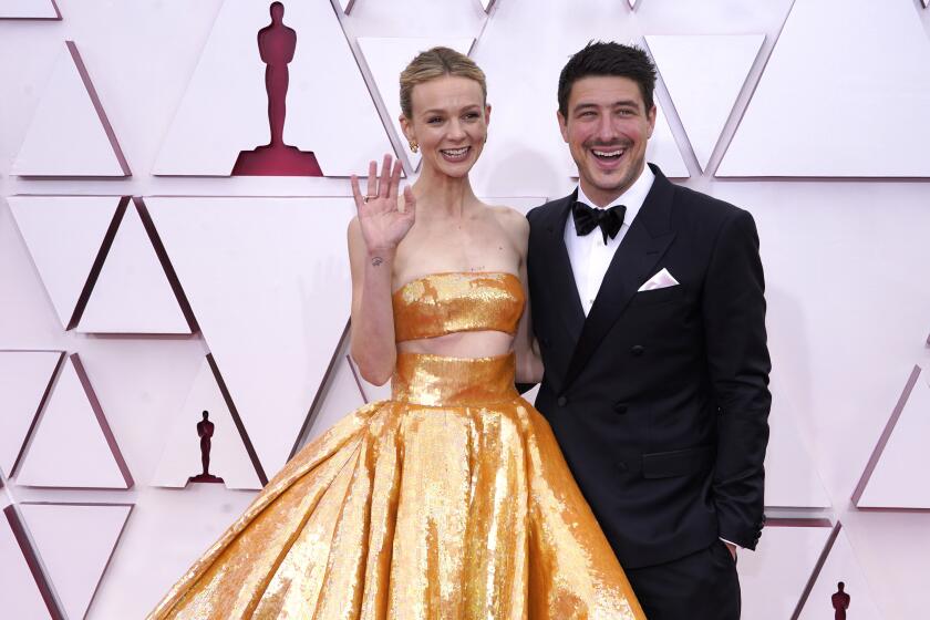 Carey Mulligan in a gold two-piece gown waving her right hand. She stands next to Marcus Mumford in a tuxedo