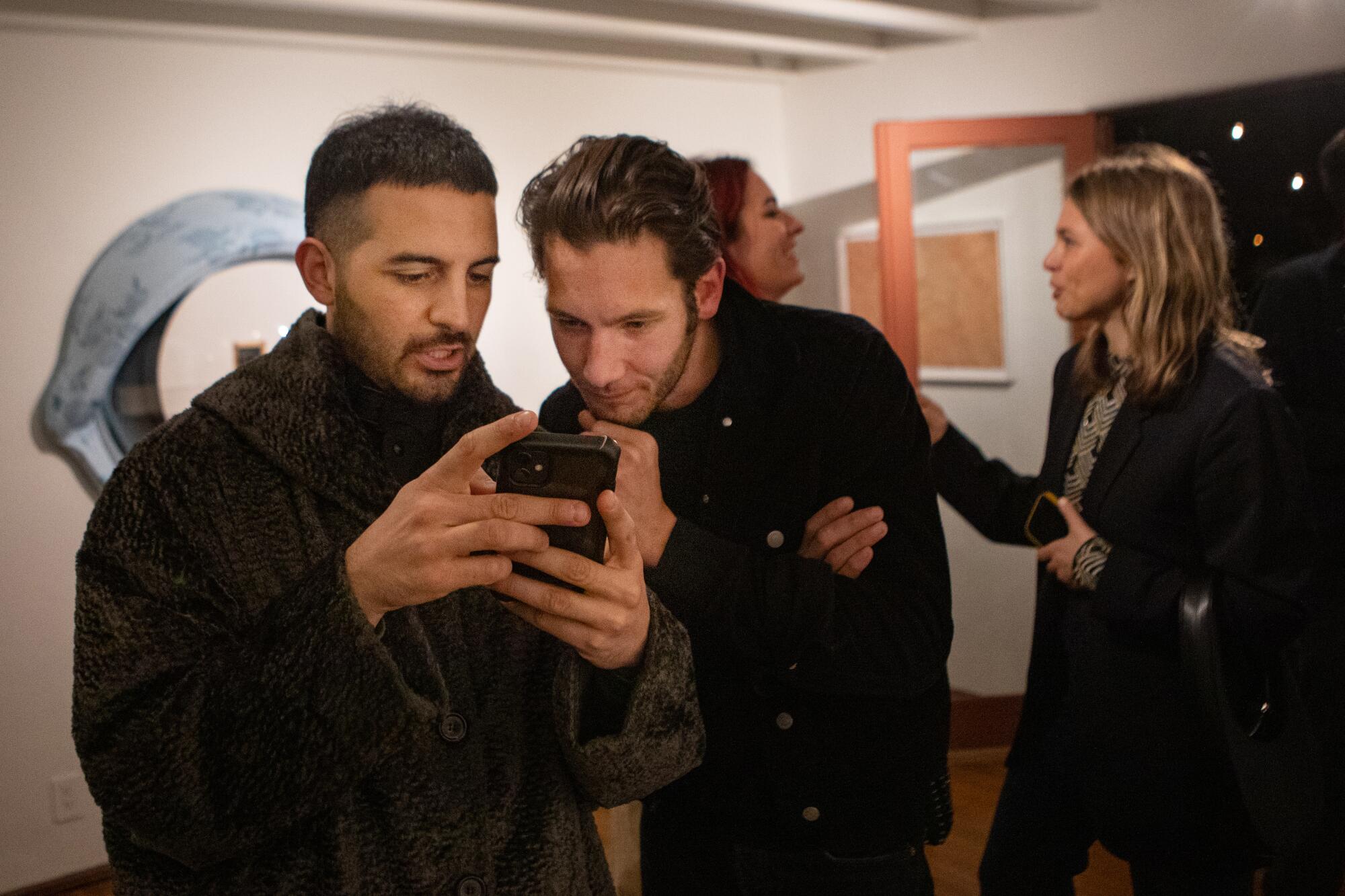 Two men in a gallery look at a phone while two women talk and laugh in the background