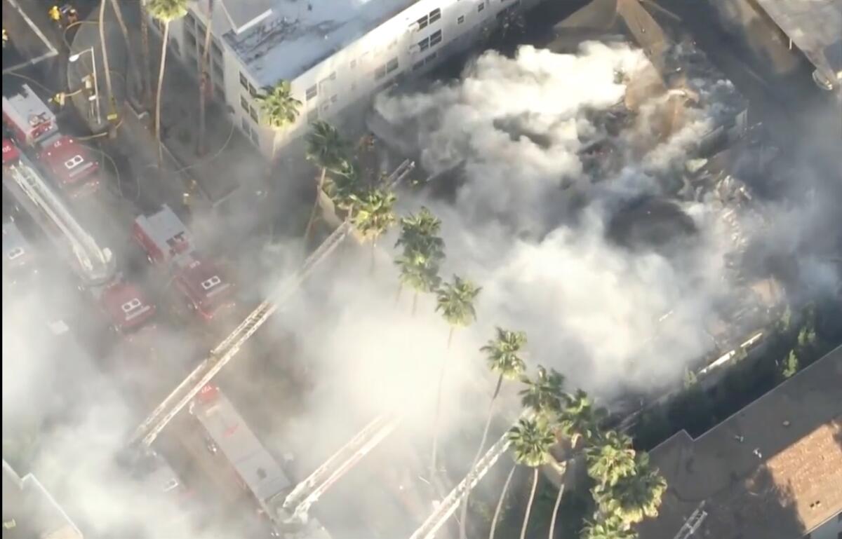An aerial view of steam and smoke rising as water is sprayed at a building on a street lined with palm trees and firetrucks