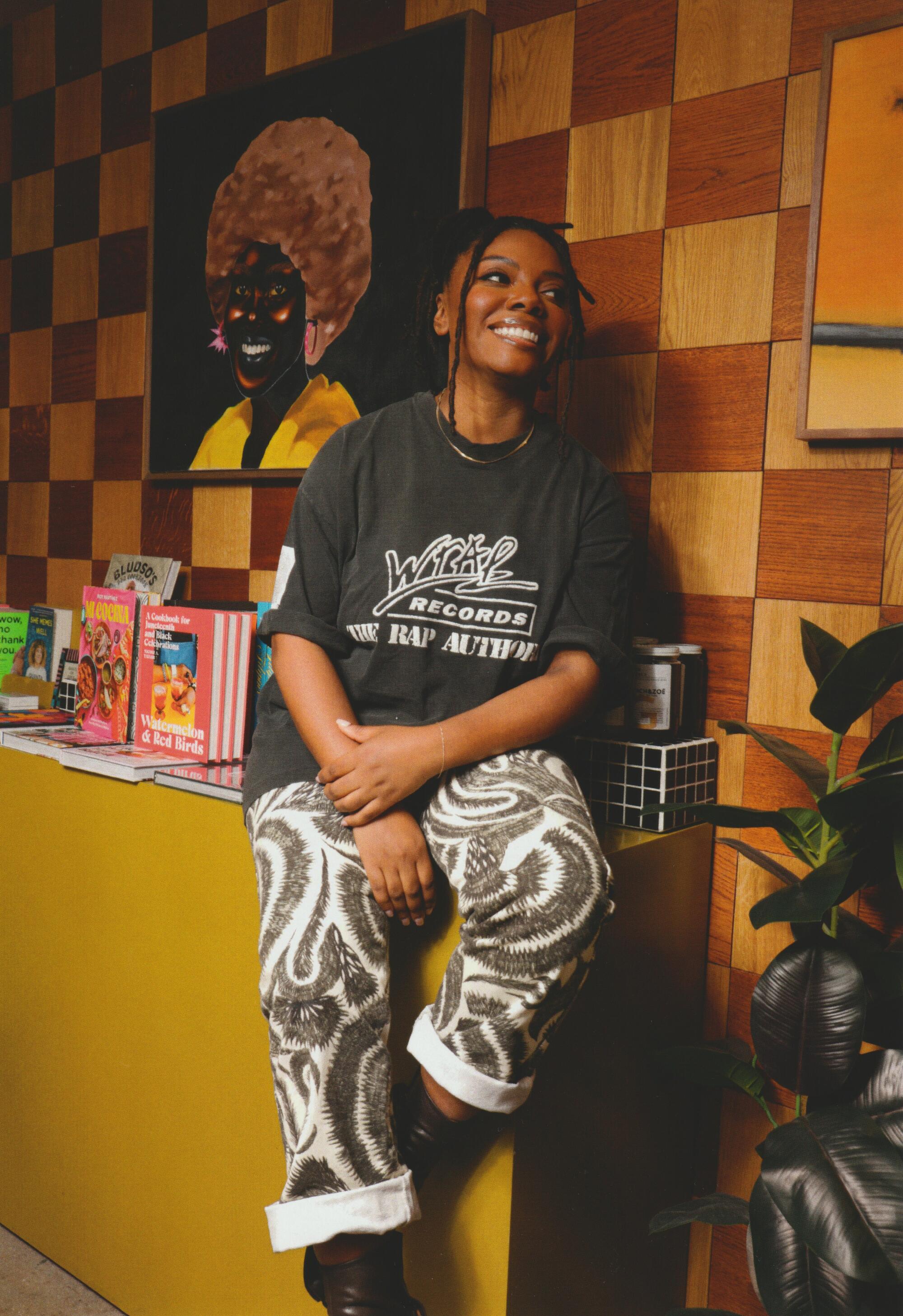 A smiling woman sits on a countertop in front of a painting and a checkerboard wood wall.