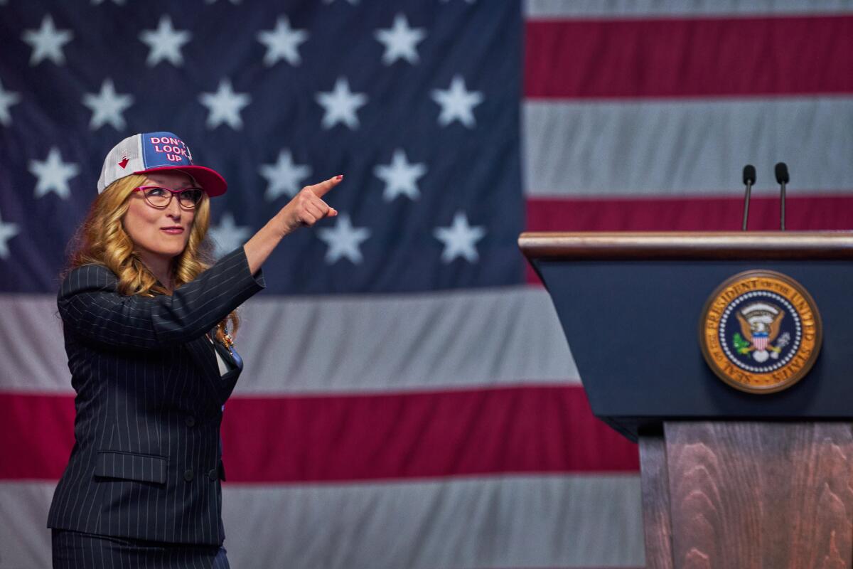 A woman in a suit and a baseball cap points in front of a giant American flag.