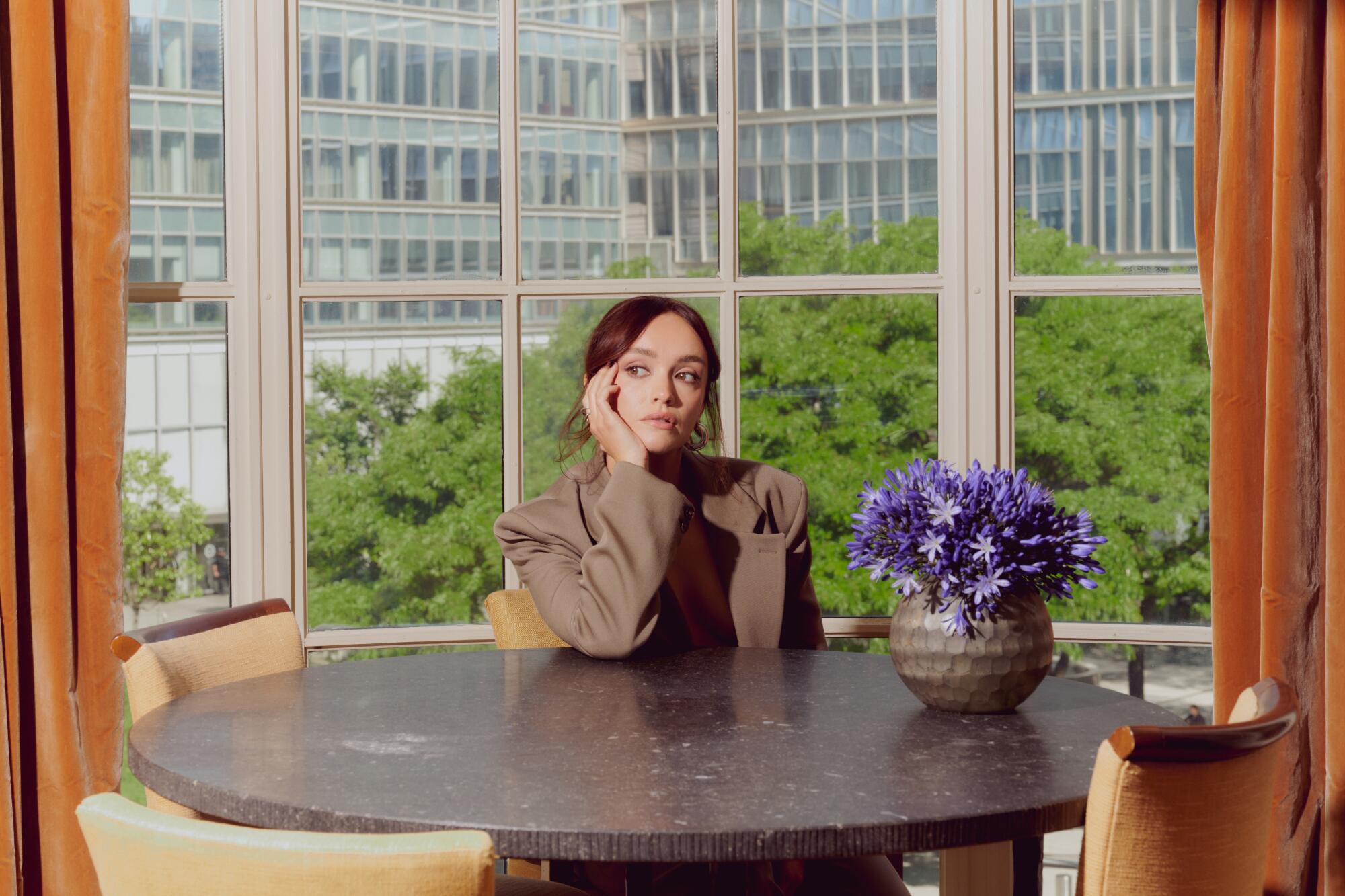 Olivia Cooke seated at a table with a flower vase next to her in front of window overlooking glass buildings.