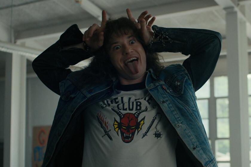 A long-haired man in a jean jacket sticks out his tongue and makes devil horns with his fingers