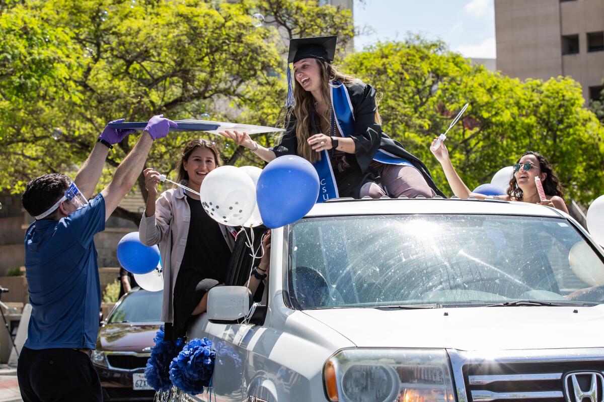 A student in graduation regalia sitting on top of a car with blue and white balloons