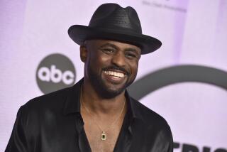 Wayne Brady smiles while wearing a black fedora and a black button up that reveals his chest and a gold necklace