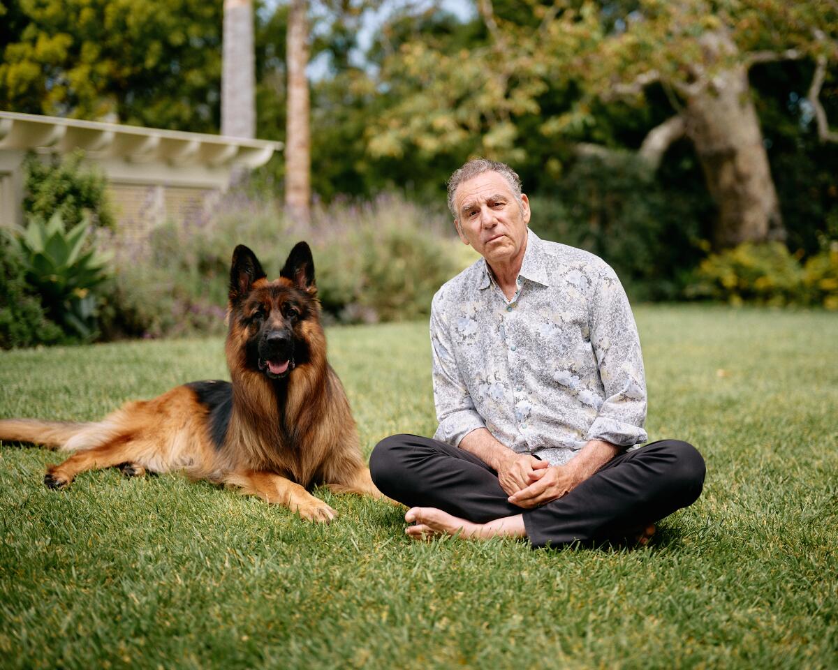 Michael Richards sits cross-legged on a lawn next to a German shepherd.