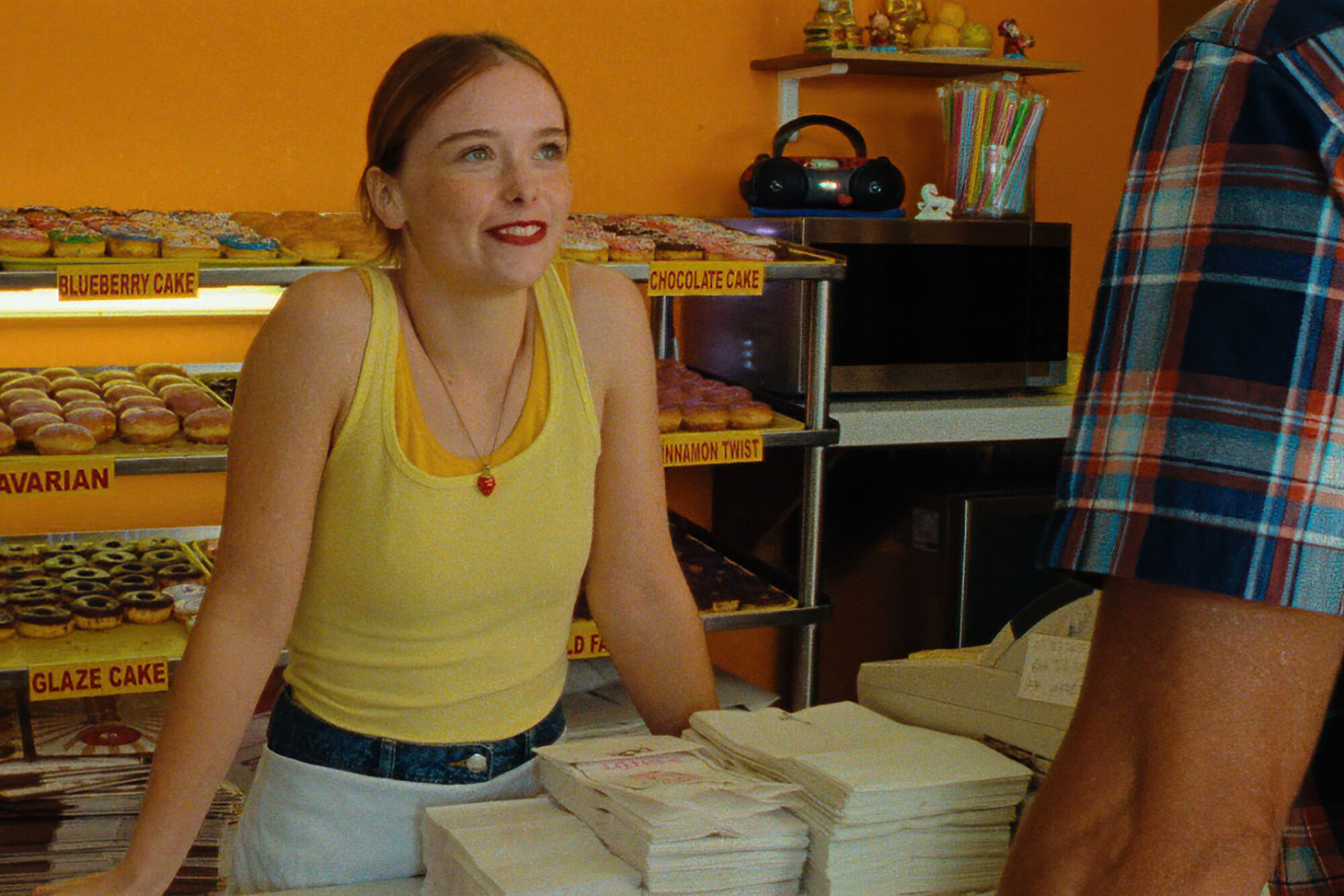 A woman in a yellow shirt smiles at a man as she stands behind the counter of a doughnut shop.